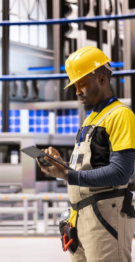 Male engineer analyzing industry 4.0 system performance in smart manufacturing plant. Black man monitoring machinery and robotic arms, ensuring optimal production and industrial automation efficiency.